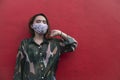 An asian woman wearing a face mask looks at the camera while posing by a red painted wall. Royalty Free Stock Photo