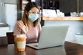 Asian woman wearing face shield and protective mask working on laptop in coffee shop