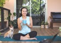 Asian woman wearing headphones, holding coffee cup, sitting  on yoga mat in balcony  with computer laptop Chihuahua dog. Yoga Royalty Free Stock Photo