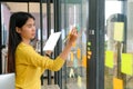 Asian woman wearing a yellow shirt with a tablet in her hand She is writing a pencil on a note on a glass wall in the office