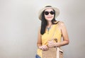 Asian woman  wearing yellow sleeveless shirt, hat and sunglasses  holding woven bag, standing on white background, smiling and Royalty Free Stock Photo