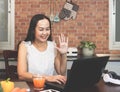 Asian woman working with computer laptop in the kitchen, using laptop for video chatting, smiling and waving to  the monitor Royalty Free Stock Photo