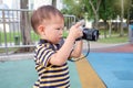 Asian 2 year old toddler boy wear camera strap taking photo using digital camera, looking at camera in park, kid photographing nat Royalty Free Stock Photo