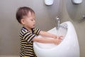 Asian 2 years old toddler baby boy child washing hands by himself on white sink and water drop from faucet in public toilet