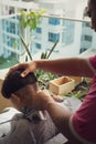 Asian young boy is getting haircut in the balcony apartment at home using electric razor. view from behind Royalty Free Stock Photo