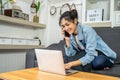 Asian young business woman is sitting on the sofa in her own house is talking on cell phone and smiling with laptop computer at Royalty Free Stock Photo