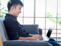 Asian young businessman typing on keyboard laptop computer while sitting on sofa couch in his apartment, freelancer working online Royalty Free Stock Photo