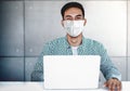 Asian Young Businessman Wearing Surgical Mask in Office. Sitting on Desk while Working on Computer Laptop. Smiling and Looking at Royalty Free Stock Photo