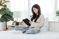 Asian young cheerful female teenage model sitting smiling on white clean sheet bed using pen with touchscreen tablet computer Royalty Free Stock Photo