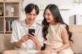 Asian young couple sit on couch or sofa looking at mobile phone smile and laughing together. Cheerful lover surfing internet and Royalty Free Stock Photo