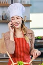 Asian young female chef housewife wears white tall cook hat and apron standing smiling on smartphone call holding wooden spoon Royalty Free Stock Photo