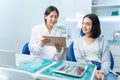 Asian young female dentist holding digital tablet and giving advice or consultation to Caucasian young girl patient. Doctor explai