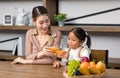 Asian young female housewife mother sitting smiling on table near bookshelf in living room at home with little girl kid daughter Royalty Free Stock Photo