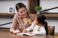 Asian young female housewife mother tutor teacher sitting smiling on table in living room at home teaching little cute Royalty Free Stock Photo