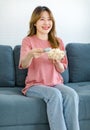 Millennial Asian young happy cheerful female housewife sitting smiling on cozy sofa holding popcorn snack glass bowl using remote Royalty Free Stock Photo