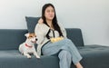 Asian young happy cheerful female owner sitting smiling on cozy sofa couch holding eating popcorn glass bowl snack watching movie