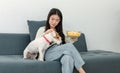 Asian young happy cheerful female owner sitting smiling on cozy sofa couch holding eating popcorn glass bowl snack watching movie