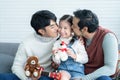 Asian young LGBTQ gay couple kissing cheek of little Caucasian adopted kid together at home. Happy daughter smiling holding dolls