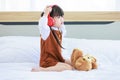Asian young lonely little cute preschooler daughter girl sitting alone on bed in bedroom holding teddy bear toy friend and red Royalty Free Stock Photo