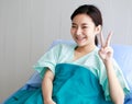 Asian young long hair happy female patient wears green hospital uniform covered by blanket sit lean on blue pillow on bed hold