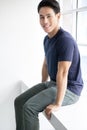 Asian young man relaxing sit on the wall hang his legs look at camera and smile. Full body portrait of handsome guy in dark blue Royalty Free Stock Photo