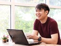 Asian young man sitting in the coffee shop with computer laptop on the table , holding mug of coffee and laughing Royalty Free Stock Photo