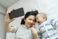 Asian Young Mother comforting or consoling her little toddler son while holding mobile phone. Newborn baby boy crying lying on bed Royalty Free Stock Photo