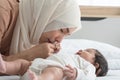 Asian young Muslim mother wearing hijab kissing hand of her newborn baby 1 month old while cute infant grabbing mom finger,