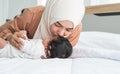 Asian young Muslim mother wearing hijab kissing her newborn baby 1 month old on forehead while cute infant sleeping on bed in Royalty Free Stock Photo