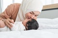 Asian young Muslim mother wearing hijab kissing her newborn baby 1 month old on forehead while cute infant sleeping on bed in Royalty Free Stock Photo