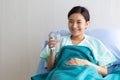 Asian young pretty female patient wears green hospital uniform sit on bed lean on blue pillow covered by blanket smiling and hold