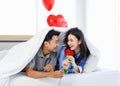 Asian young romantic lover couple male boyfriend giving red rose surprising female girlfriend laying down under blanket on bed