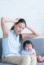 Asian young stressed mother holding head with hands sitting with 7 months crying pitiful infant baby sitting on sofa at home.