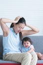 Asian young stressed mother holding head with hands sitting with 7 months crying pitiful infant baby sitting on sofa at home.