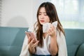 woman sick sitting down on sofa with cold blowing her nose and checking smart mobile phone Royalty Free Stock Photo