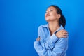 Asian young woman standing over blue background hugging oneself happy and positive, smiling confident Royalty Free Stock Photo