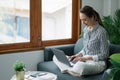 Asian young woman working from home office sit on couch hold paper invoice check information online Royalty Free Stock Photo