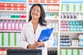 Asian young woman working at pharmacy drugstore holding clipboard looking positive and happy standing and smiling with a confident Royalty Free Stock Photo