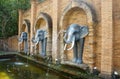Replica of Asian elephants with long white tusks and a water-spouting trunk on the brick wall. Fountain in the park.