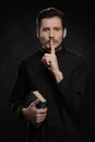 Asking to keep silence. Portrait of priest holding his hand against lips while standing against black background