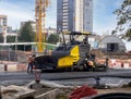 Asphalting of the road by an asphalt paver at a city construction site. Construction materials are stacked next to the road.