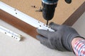 Assembling furniture, gloved hands install roller guides on box using cordless screwdriver, close-up