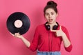 Astonish young charming girl holding music plate in hand and pointing at it with forefinger, keeps mouth opened, sees vintage disc Royalty Free Stock Photo