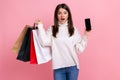 Astonished dark haired female posing with shopping bags, showing phone with blank screen for ad.