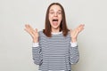 Astonished excited amazed woman with brown hair wearing striped shirt standing isolated over gray background raised her arms