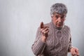 An astonished handsome elderly man with gray hair dressed in sweater standing near white wall pointing with finger wanting to say