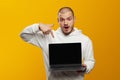 Astonished hipster man wearing white hoodie holding laptop in hands while pointing at screen Royalty Free Stock Photo