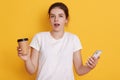 Astonished lady with opened mouth standing against yellow background, holding smart phone and take away coffee in hands, wearing Royalty Free Stock Photo