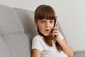Astonished little girl wearing white t shirt sitting on sofa at home, holding smart phone in hands, talking with friend, hearing Royalty Free Stock Photo