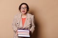Astonished woman school teacher holds heavy volumes of hardcover books, expresses stupefaction at camera, cream backdrop Royalty Free Stock Photo